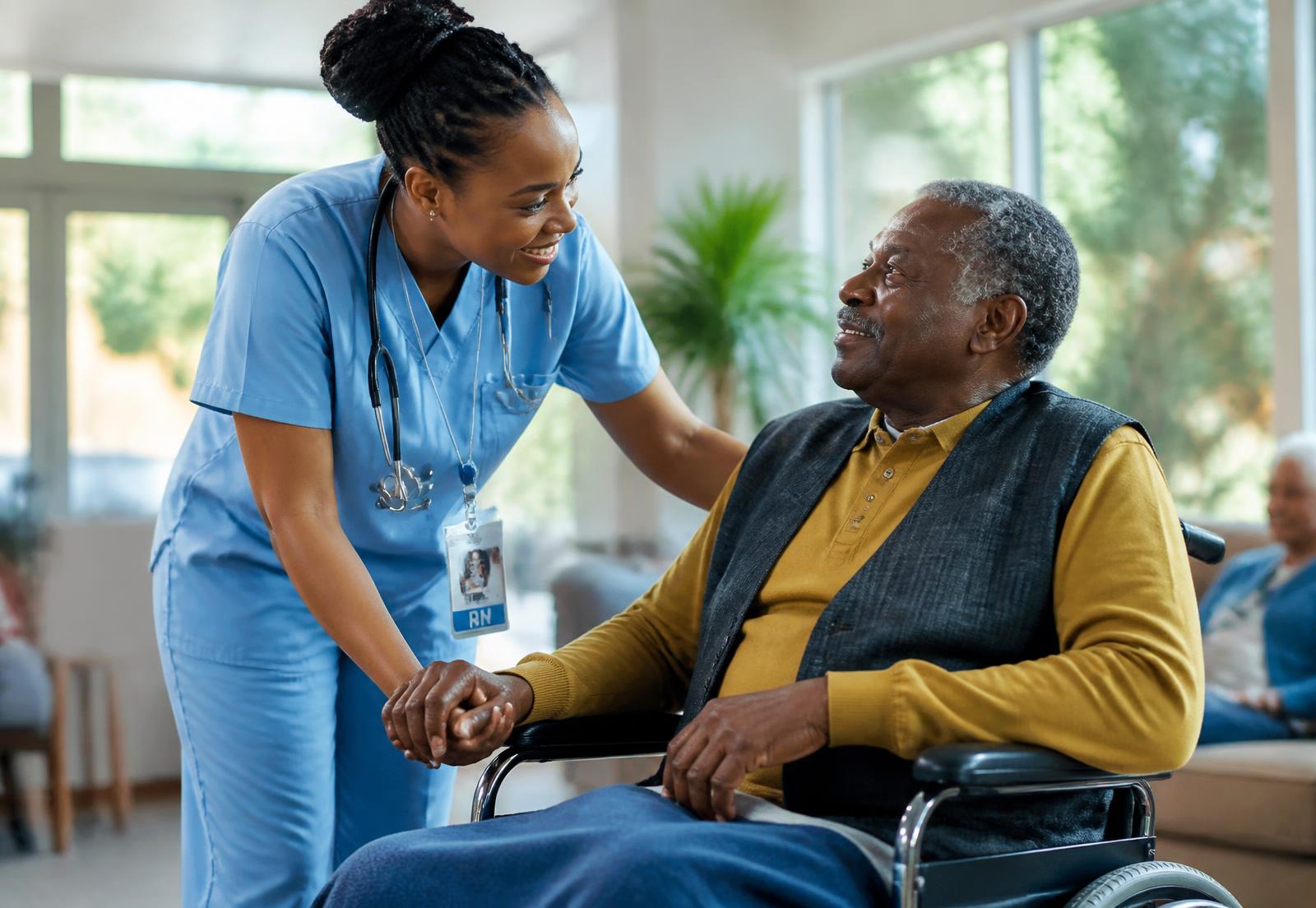 A nurse in scrubs holding the hand of a smiling older resident seated in a wheelchair in a sunlit senior living common room